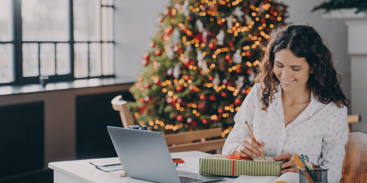 Frau sitzt an einem Tisch und beschriftet ein Weihnachtspäckchen. Im Hintergrund ein beleuchteter Weihnachtsbaum.